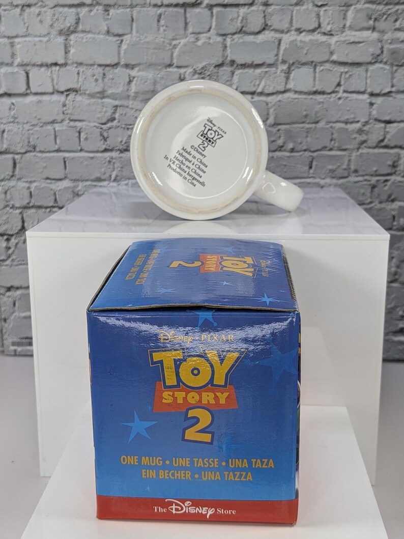 May include: A white ceramic mug with a handle, positioned behind its blue cardboard packaging box. The mug base features the Toy Story 2 logo and manufacturing text. The box displays the Toy Story 2 logo, blue stars, and the text One Mug.