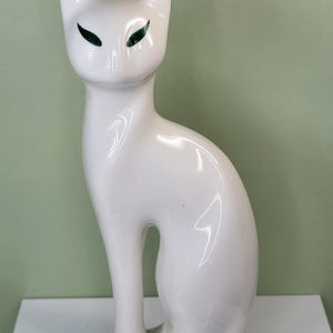 May include: A white ceramic cat figurine with green eyes. The cat is in a seated position with its tail curled. The figurine has a glossy finish and is displayed on a white surface against a light green background.