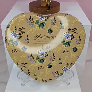 May include: A heart-shaped tin with a floral design in shades of gold, blue, and green. The tin features the brand name "Brighton" and text indicating it's jewelry packaging. The tin is displayed on a clear stand.