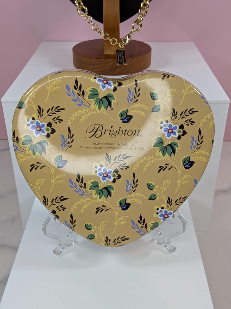 May include: A heart-shaped tin with a floral design in shades of gold, blue, and green. The tin features the brand name "Brighton" and text indicating it's jewelry packaging. The tin is displayed on a clear stand.