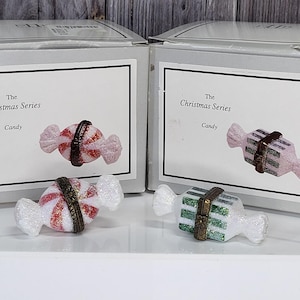 May include: Two boxes and two candy-shaped ornaments. The boxes have a label that reads "The Christmas Series Candy". The candy ornaments are white, red, and green with a gold-colored band around the middle. The ornaments are on a white surface.