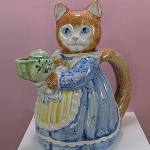 May include: A ceramic teapot shaped like a cat, with orange and white fur, blue eyes, and a blue dress adorned with white floral patterns. The cat holds a green teapot with floral designs. The handle is the cat's tail.