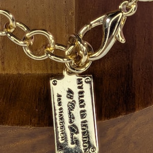 May include: Close-up of a gold-colored necklace with a lobster clasp and a rectangular tag. The tag is engraved with the text "MY FLAT IN LONDON 49 Black Place" and "Jewellery Handmade". The chain links are large and oval-shaped.