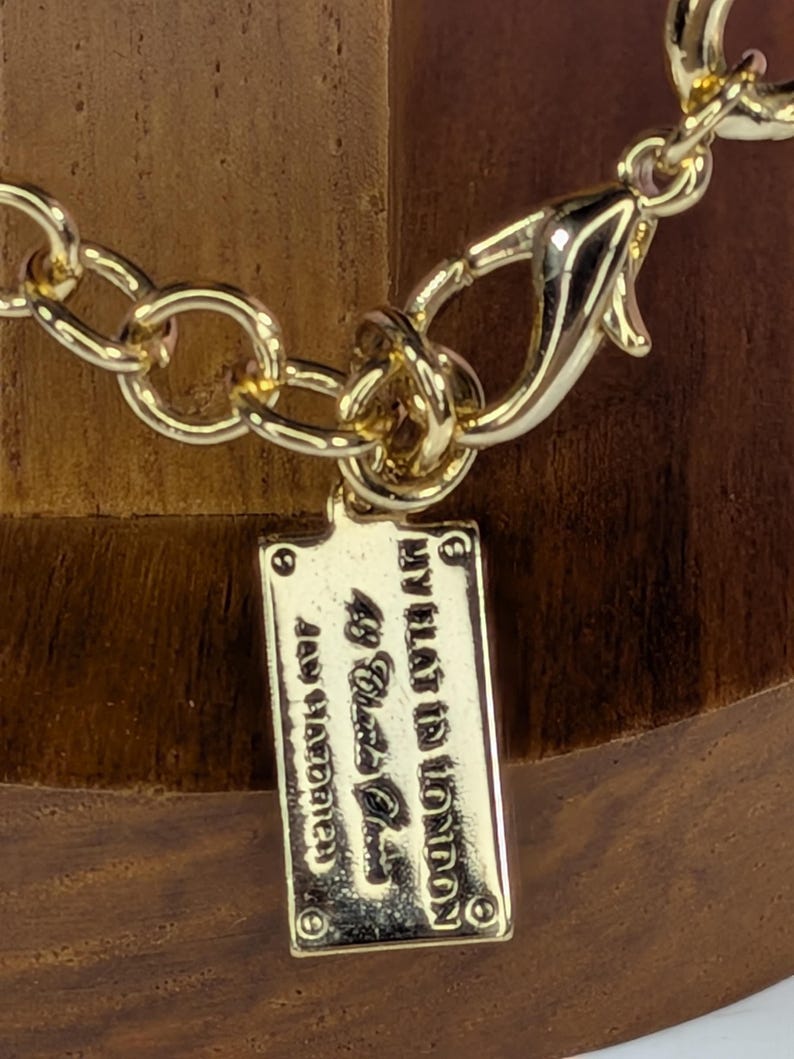 May include: Close-up of a gold-colored necklace with a lobster clasp and a rectangular tag. The tag is engraved with the text "MY FLAT IN LONDON 49 Black Place" and "Jewellery Handmade". The chain links are large and oval-shaped.