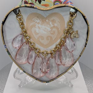 May include: A gold-toned chain necklace with large, pink teardrop-shaped pendants. The necklace is displayed inside a heart-shaped box with the word "Brighton" visible. A small, jeweled dog charm hangs from the chain.