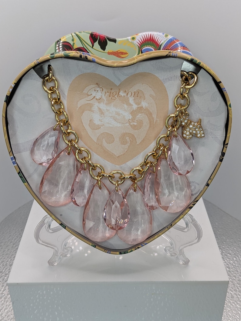 May include: A gold-toned chain necklace with large, pink teardrop-shaped pendants. The necklace is displayed inside a heart-shaped box with the word "Brighton" visible. A small, jeweled dog charm hangs from the chain.
