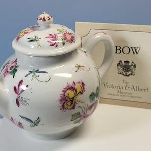 May include: White porcelain teapot with a lid, decorated with colorful floral and dragonfly designs. The teapot has a curved spout and handle. A card with the text "BOW" and "The Victoria & Albert Museum" is next to the teapot.