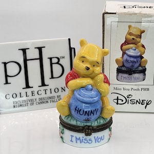 May include: A Winnie the Pooh figurine, featuring the character in a red shirt, yellow fur, and a blue honey pot. The figurine is on a decorative box with the text "I Miss You" and "Hunny". A white PHB Collection sign is on the left.