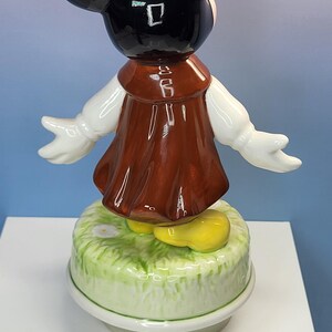 May include: A ceramic figurine of a cartoon character with black ears, a brown dress, white gloves, and yellow shoes. The figurine is standing on a green and white base with a floral design.