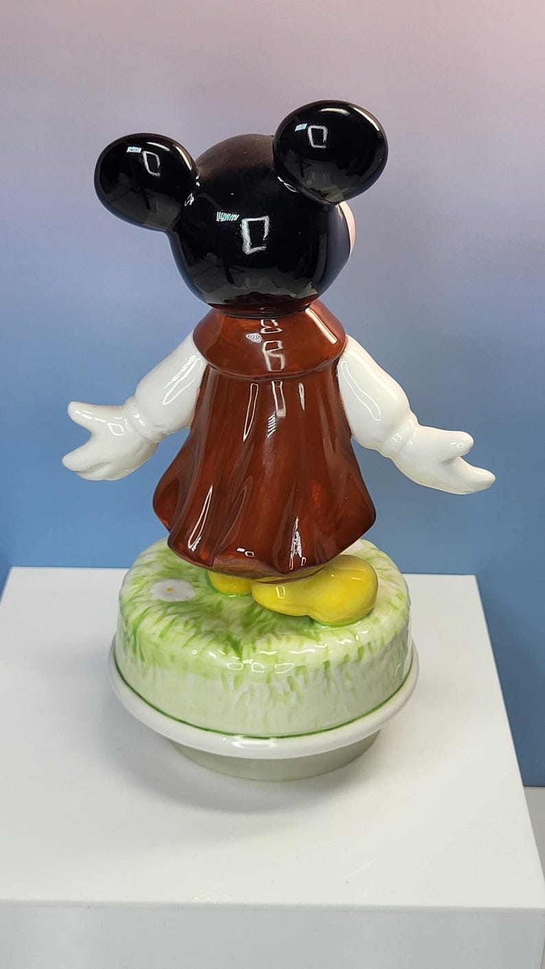 May include: A ceramic figurine of a cartoon character with black ears, a brown dress, white gloves, and yellow shoes. The figurine is standing on a green and white base with a floral design.