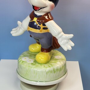 May include: A ceramic Mickey Mouse figurine with outstretched arms, wearing a brown vest, blue pants, and yellow shoes. The figurine stands on a green base with white flowers. The figure has black ears and a smiling face.
