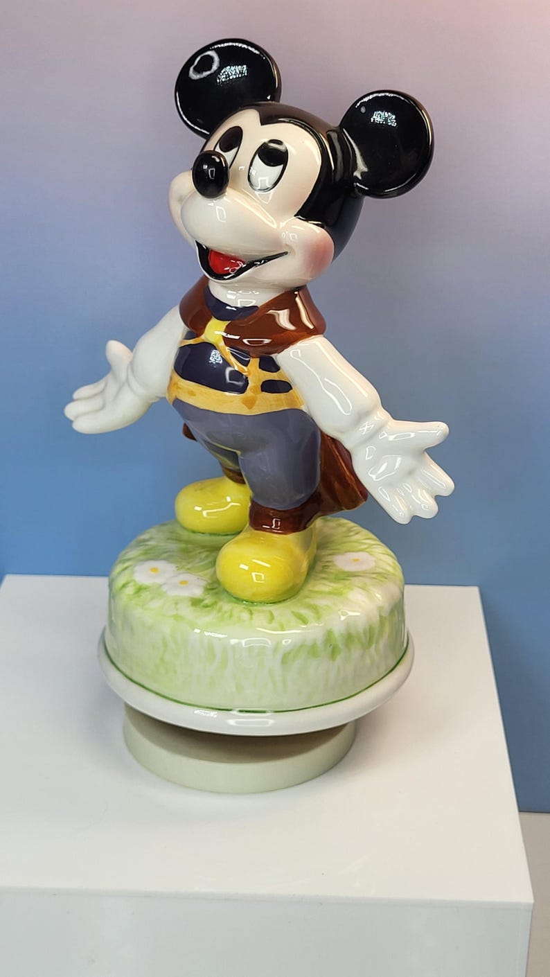 May include: A ceramic Mickey Mouse figurine with outstretched arms, wearing a brown vest, blue pants, and yellow shoes. The figurine stands on a green base with white flowers. The figure has black ears and a smiling face.