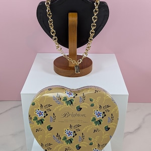 May include: A gold-toned chain necklace displayed on a black jewelry bust. A heart-shaped tin with a floral pattern and the Brighton logo sits below. The tin is gold with blue and white flowers.