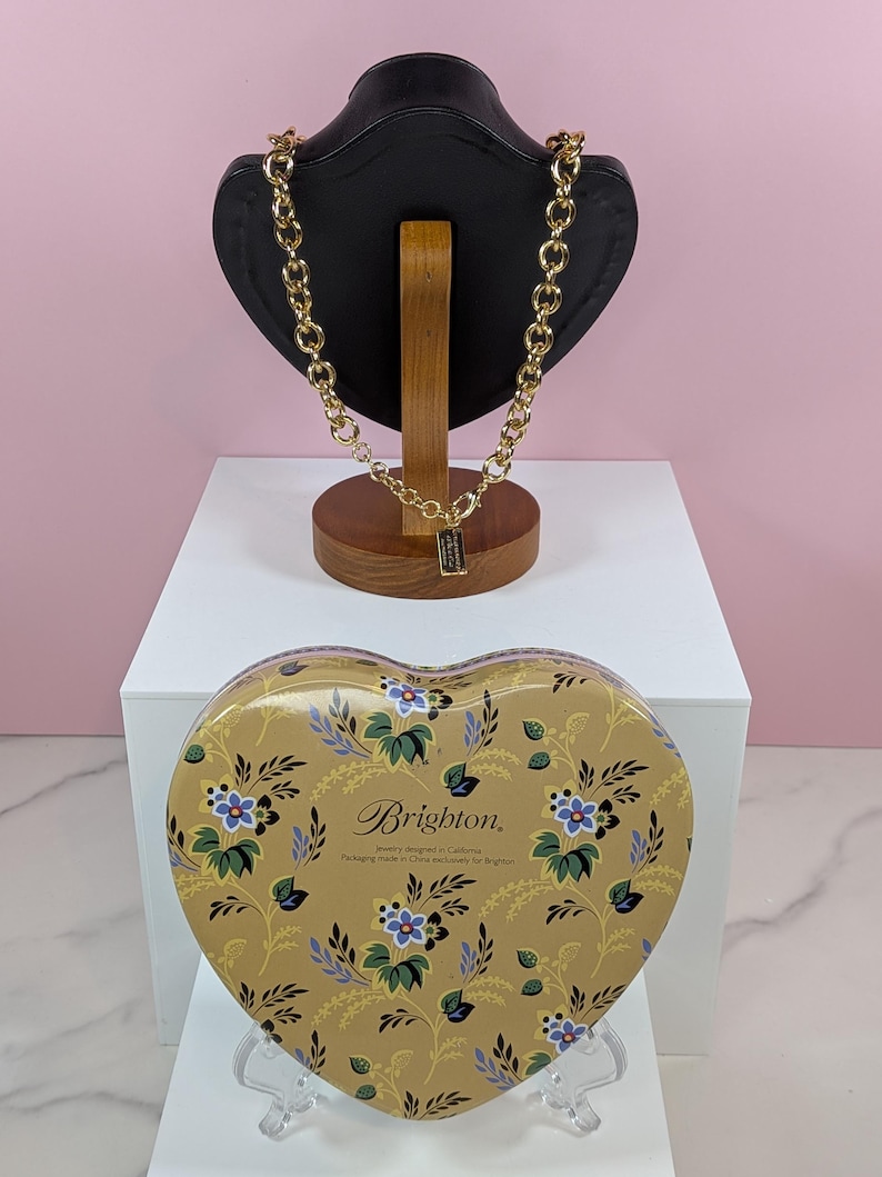 May include: A gold-toned chain necklace displayed on a black jewelry bust. A heart-shaped tin with a floral pattern and the Brighton logo sits below. The tin is gold with blue and white flowers.