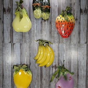 May include: A collection of six ceramic fruit and vegetable wall plaques featuring a glossy yellow pear, two dark green cucumbers, a red strawberry, a bunch of yellow bananas, a yellow lemon, and a purple eggplant, each topped with a green ribbon.