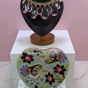May include: A gold-toned necklace with large, pink teardrop-shaped pendants is displayed on a black bust. Below, a heart-shaped tin with a floral design in green, red, blue, and black sits on a clear stand. The tin has the word "Brighton" on it.