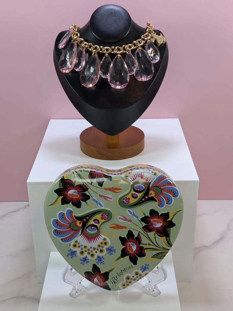 May include: A gold-toned necklace with large, pink teardrop-shaped pendants is displayed on a black bust. Below, a heart-shaped tin with a floral design in green, red, blue, and black sits on a clear stand. The tin has the word "Brighton" on it.