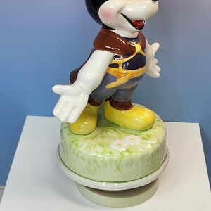 May include: A ceramic Mickey Mouse figurine, standing on a green base with white daisies. Mickey is wearing a brown top, yellow shoes, and dark blue pants. The figurine has black ears and a smiling expression.
