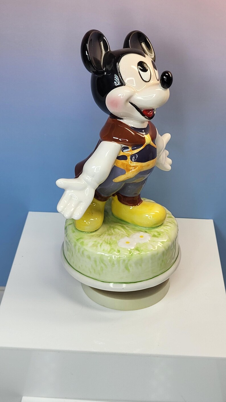 May include: A ceramic Mickey Mouse figurine, standing on a green base with white daisies. Mickey is wearing a brown top, yellow shoes, and dark blue pants. The figurine has black ears and a smiling expression.