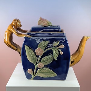 May include: A ceramic teapot with a dark blue body, adorned with a floral design and a monkey handle. The spout and handle are a golden-brown color. The lid features a pink and white flower. The teapot is displayed on a white surface.