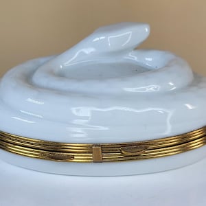 May include: A white ceramic trinket box with a coiled snake design on the lid. The box has a gold-colored metal band around the base. The snake's head is raised, adding a three-dimensional element to the design.