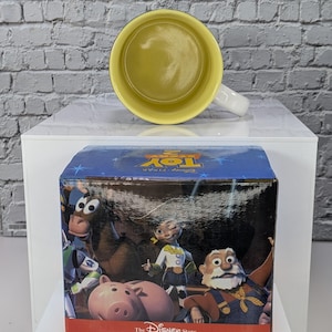 May include: A ceramic mug with a bright yellow interior and a white exterior, positioned behind a square cardboard box. The box features a blue top with the text Toy Story 2 and a front panel displaying characters including Jessie, Bullseye, Hamm, and Stinky Pete, with the text The Disney Store at the bottom.