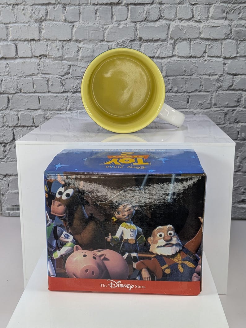 May include: A ceramic mug with a bright yellow interior and a white exterior, positioned behind a square cardboard box. The box features a blue top with the text Toy Story 2 and a front panel displaying characters including Jessie, Bullseye, Hamm, and Stinky Pete, with the text The Disney Store at the bottom.