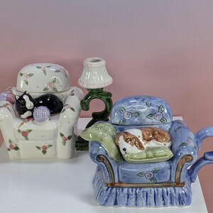 May include: Two ceramic armchair-shaped trinket boxes and a small lamp figurine. One white chair features a black cat and pink floral patterns. The blue chair features a brown and white dog on a green cushion. A dark green lamp stands between them.