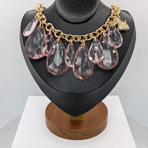 May include: A gold-tone chain necklace with large, faceted, pink teardrop pendants. The necklace is displayed on a black jewelry bust. A small, rhinestone-studded dog charm hangs from the chain.