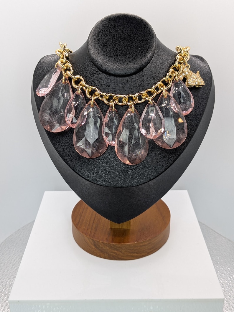 May include: A gold-tone chain necklace with large, faceted, pink teardrop pendants. The necklace is displayed on a black jewelry bust. A small, rhinestone-studded dog charm hangs from the chain.