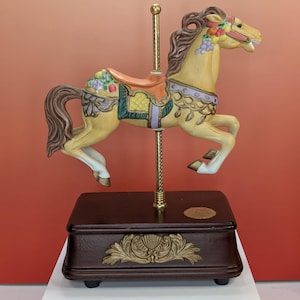 May include: A decorative carousel horse figurine in a leaping pose. The horse is painted yellow with a brown mane and tail, adorned with floral and fruit accents. It sits atop a dark brown music box base with gold-colored embellishments.