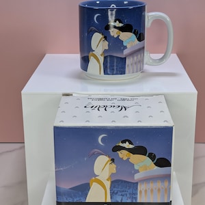 May include: A blue ceramic mug featuring a scene from Aladdin, with Aladdin and Jasmine under a crescent moon. The mug has a white handle and is displayed with its matching box, which also has the same design.