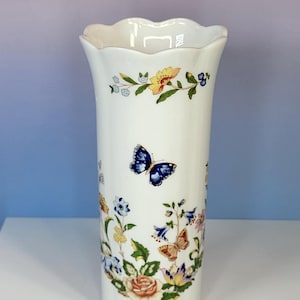 May include: White ceramic vase with a scalloped rim, adorned with colorful floral and butterfly designs. The vase features a cylindrical shape and is decorated with blue, yellow, and pink flowers, along with blue butterflies.