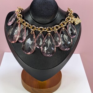 May include: A gold-toned chain necklace with large, clear, pink teardrop-shaped pendants. The necklace is displayed on a black jewelry bust. A small, gold-toned charm with clear crystals is attached to the chain.