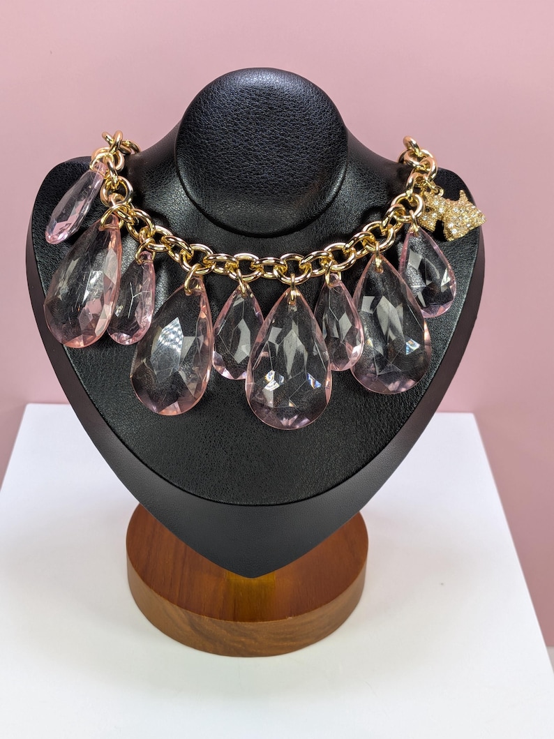 May include: A gold-toned chain necklace with large, clear, pink teardrop-shaped pendants. The necklace is displayed on a black jewelry bust. A small, gold-toned charm with clear crystals is attached to the chain.