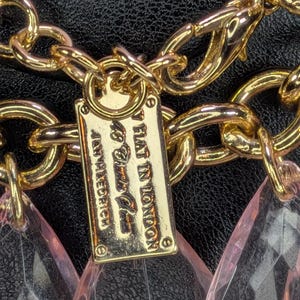 May include: Gold-toned chain necklace with a rectangular tag that reads "BY FLAT IN LONDON" and pink teardrop-shaped pendants. The necklace is set against a black background.