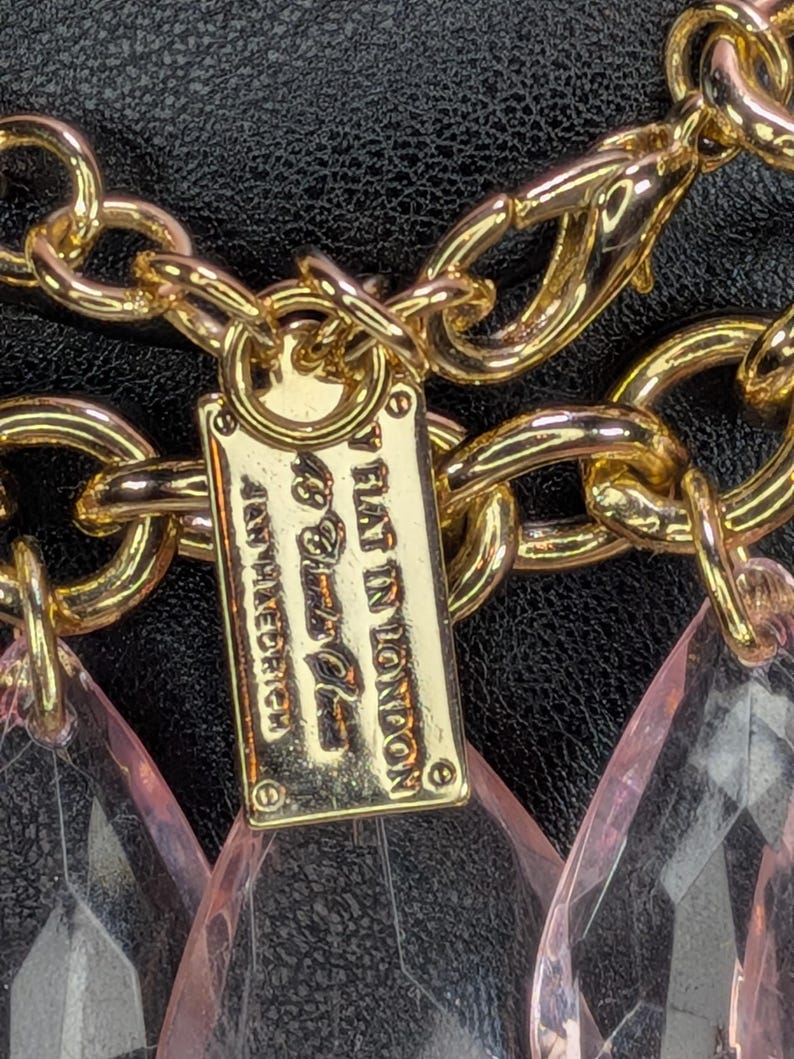 May include: Gold-toned chain necklace with a rectangular tag that reads "BY FLAT IN LONDON" and pink teardrop-shaped pendants. The necklace is set against a black background.
