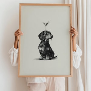 May include: A framed print of a black and brown dachshund with a martini glass balanced on its head. The martini glass contains an olive. The artwork is in monochrome, with a light beige background and a natural wood frame.