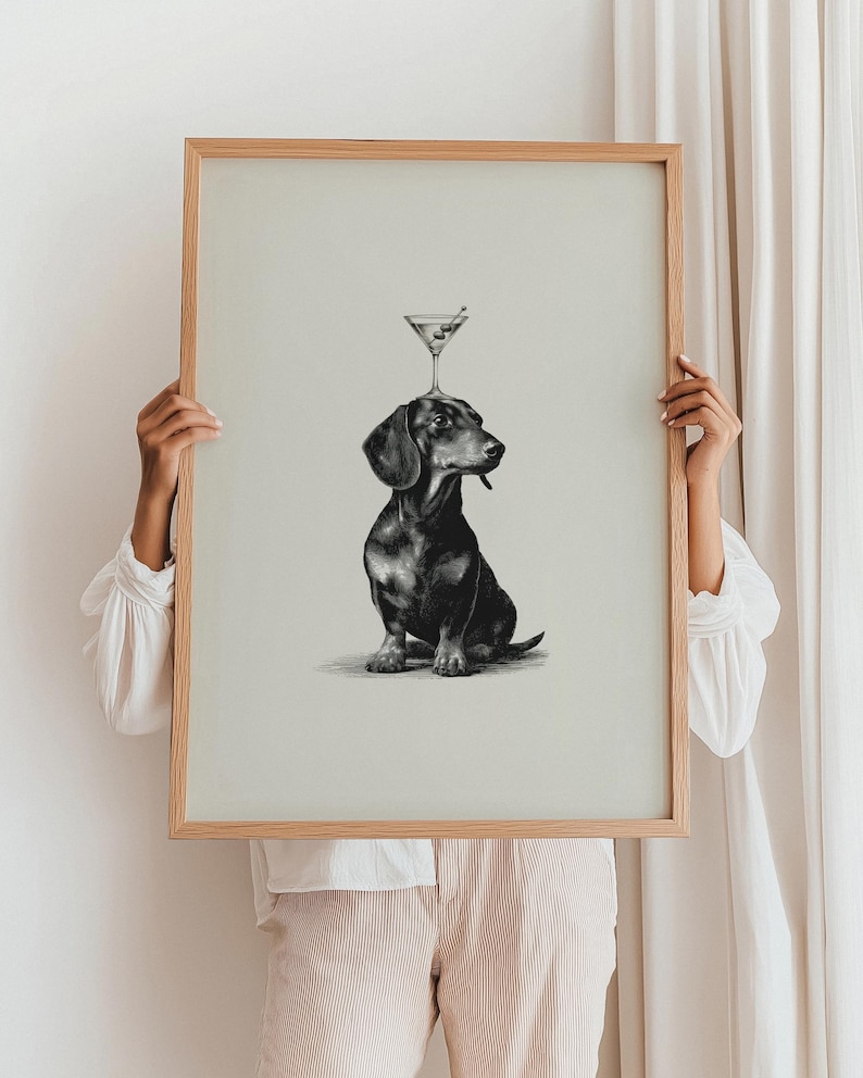 May include: A framed print of a black and brown dachshund with a martini glass balanced on its head. The martini glass contains an olive. The artwork is in monochrome, with a light beige background and a natural wood frame.