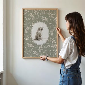 May include: A framed print with a dog portrait in an oval, set against a sage green floral design. The wooden frame complements the artwork. The print is being held up against a white wall.