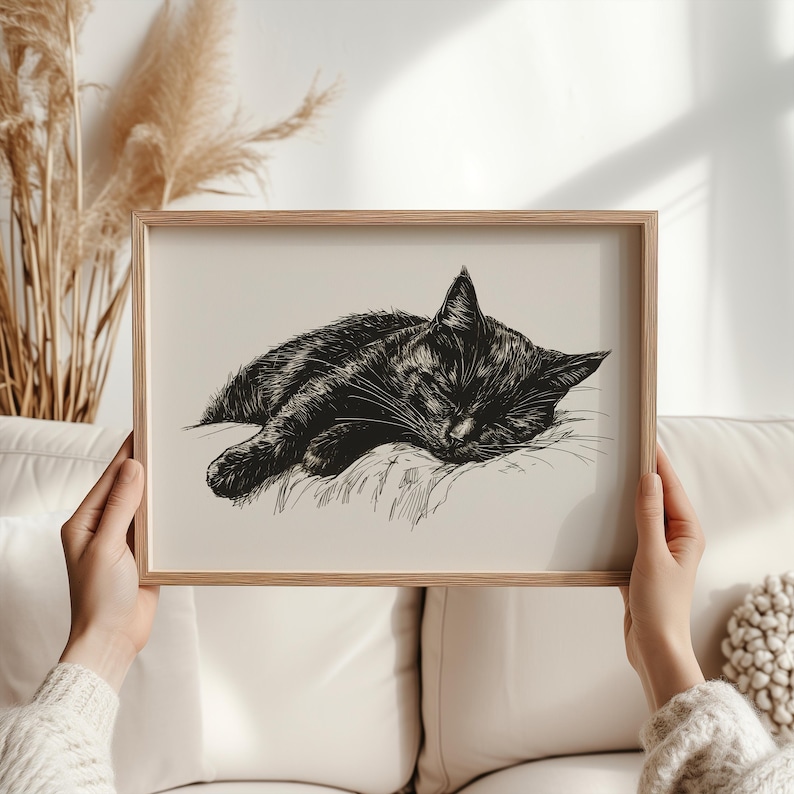 May include: Black and white line art print of a cat sleeping on a pillow. The print is framed in a light wood frame.