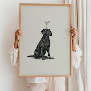 May include: A framed print featuring a black Labrador Retriever with a martini glass balanced on its head. The artwork is in grayscale, with a light background and a natural wood frame. The dog is sitting, looking to the side.