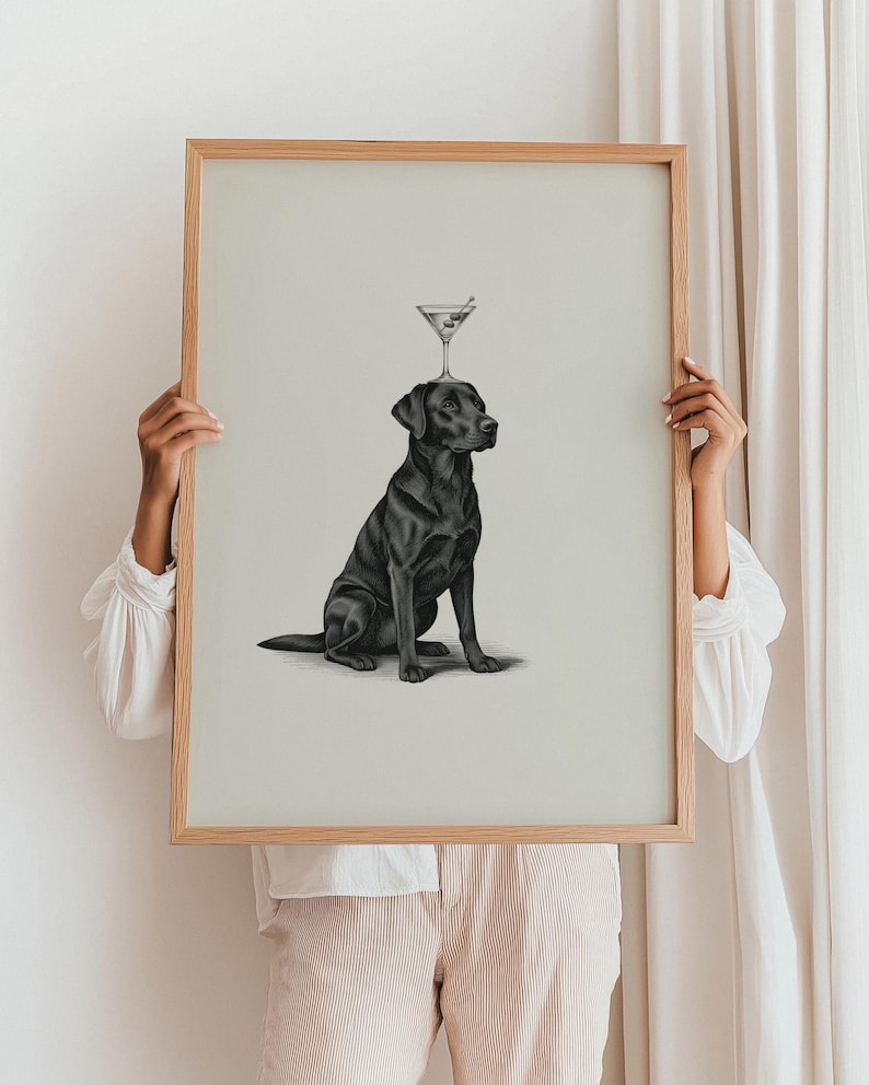 May include: A framed print featuring a black Labrador Retriever with a martini glass balanced on its head. The artwork is in grayscale, with a light background and a natural wood frame. The dog is sitting, looking to the side.