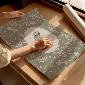 May include: A poster with a dog illustration in an oval frame, set against a sage green backdrop with a white floral design. The poster is displayed on a wooden surface. The overall aesthetic is delicate and artistic.