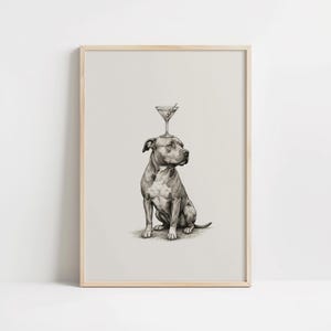 May include: A framed black and white print of a seated dog with a martini glass balanced on its head. The martini glass contains a green olive. The dog is a pit bull terrier, and the print has a light beige background.