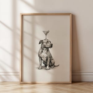 May include: A framed print featuring a detailed pencil drawing of a seated dog with a martini glass balanced on its head. The artwork is in grayscale, with a light beige background and a natural wood frame. The dog is a pit bull type breed.