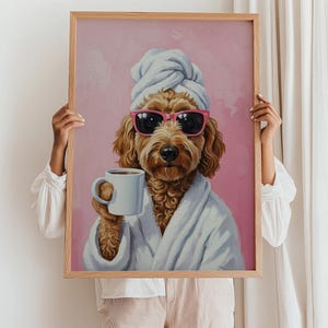以下が含まれることがあります： サングラス、タオル、白いバスローブを着てコーヒーカップを持った犬の額装プリント。犬は茶色で、背景はピンク色です。フレームはライトブラウンです。