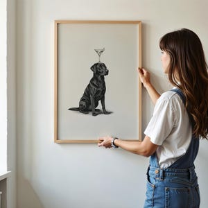 May include: A framed print featuring a black Labrador Retriever with a martini glass balanced on its head. The artwork is in a minimalist style, with the dog in a seated position. The frame is made of light-colored wood.
