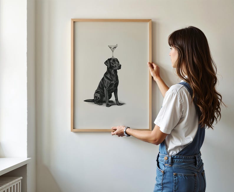 May include: A framed print featuring a black Labrador Retriever with a martini glass balanced on its head. The artwork is in a minimalist style, with the dog in a seated position. The frame is made of light-colored wood.