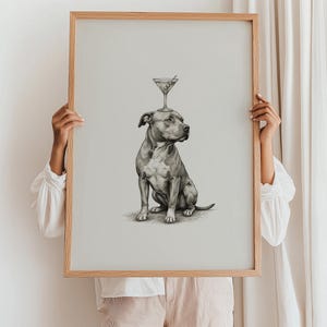May include: A framed print featuring a detailed pencil drawing of a seated pit bull with a martini glass balanced on its head. The artwork is in grayscale, with a light wooden frame. The background is a neutral, off-white color.